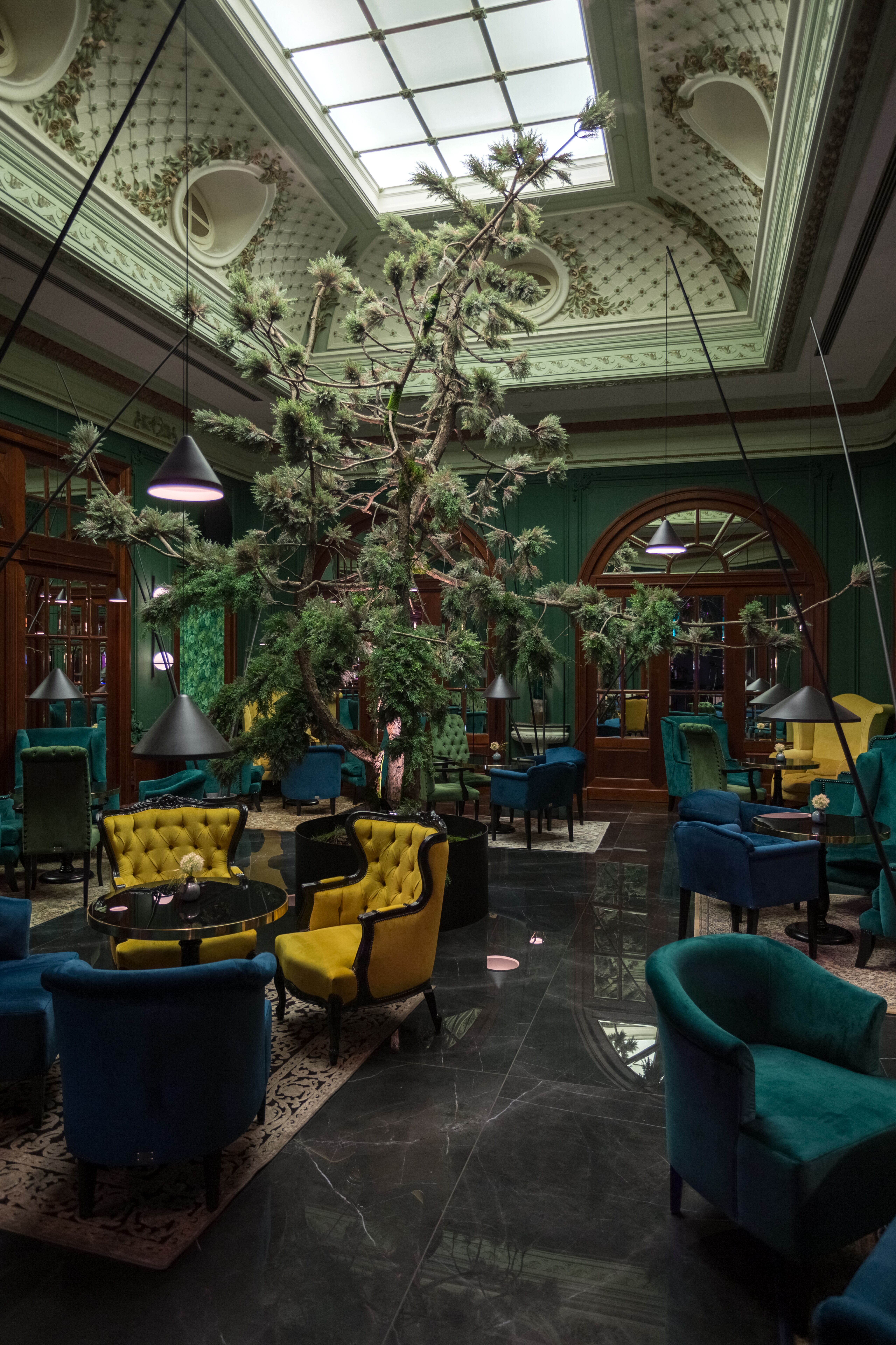 Interiér baru v zelených farbách so žltým akcentom stoličiek a dominantným stromov v strede fotky.