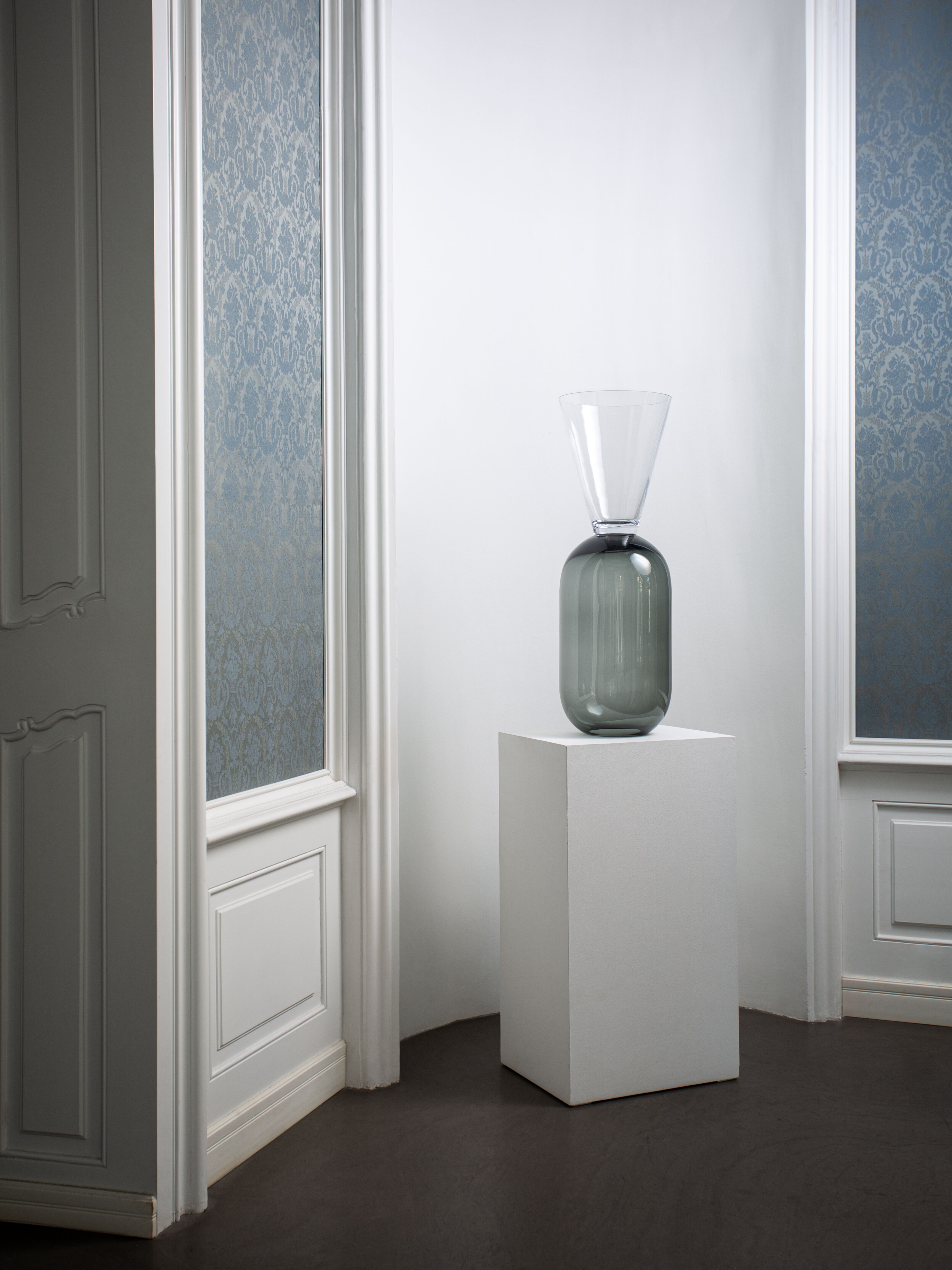 Veľká šedo číra váza zo skla na podstavci zakomponovaná do bieleho výklenku v interiéri. 