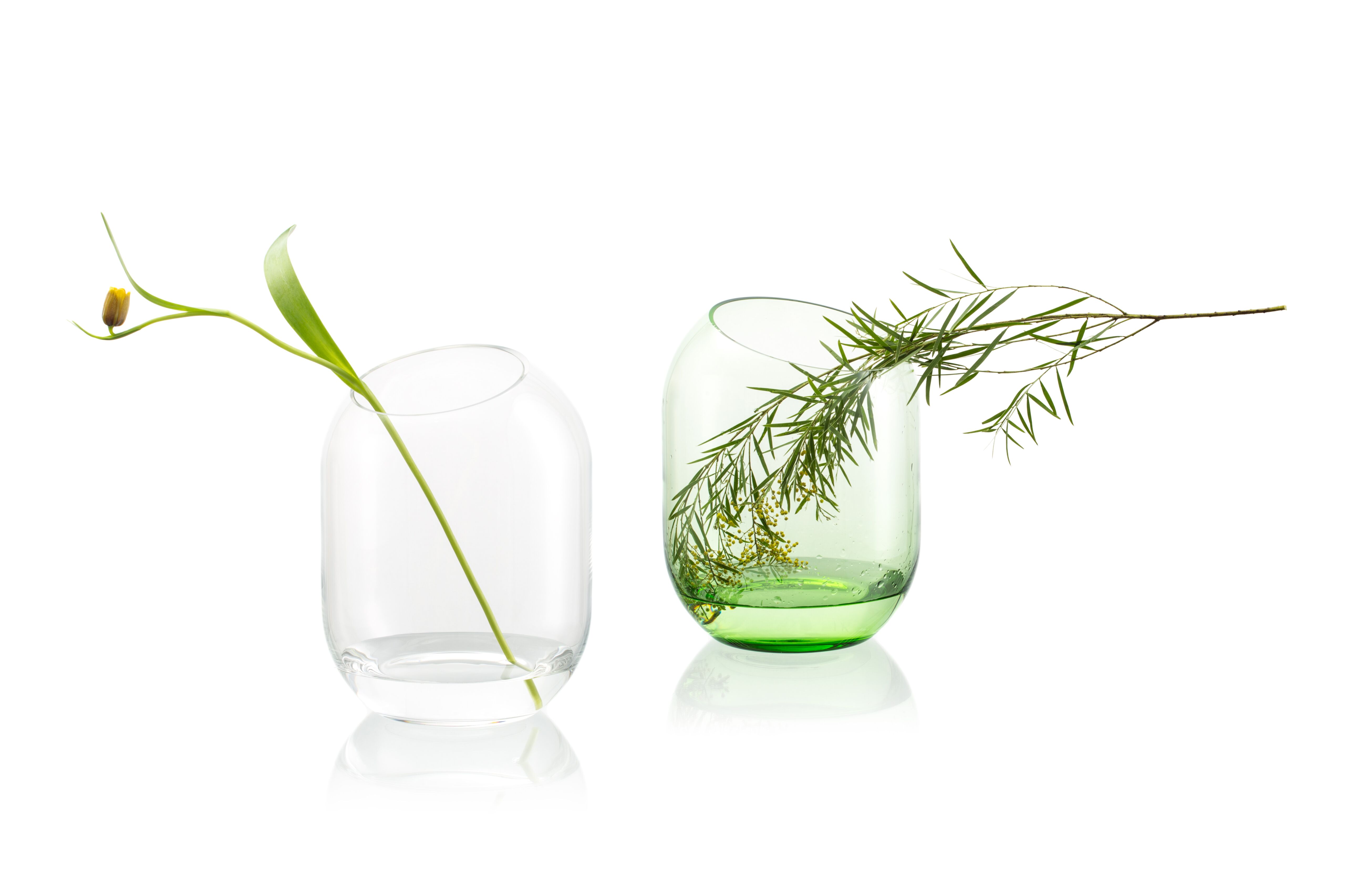 Dve sklenené vázy s kvetmi stojace vedľa seba, jedna číra a druhá zelena na bielom pozadí. 